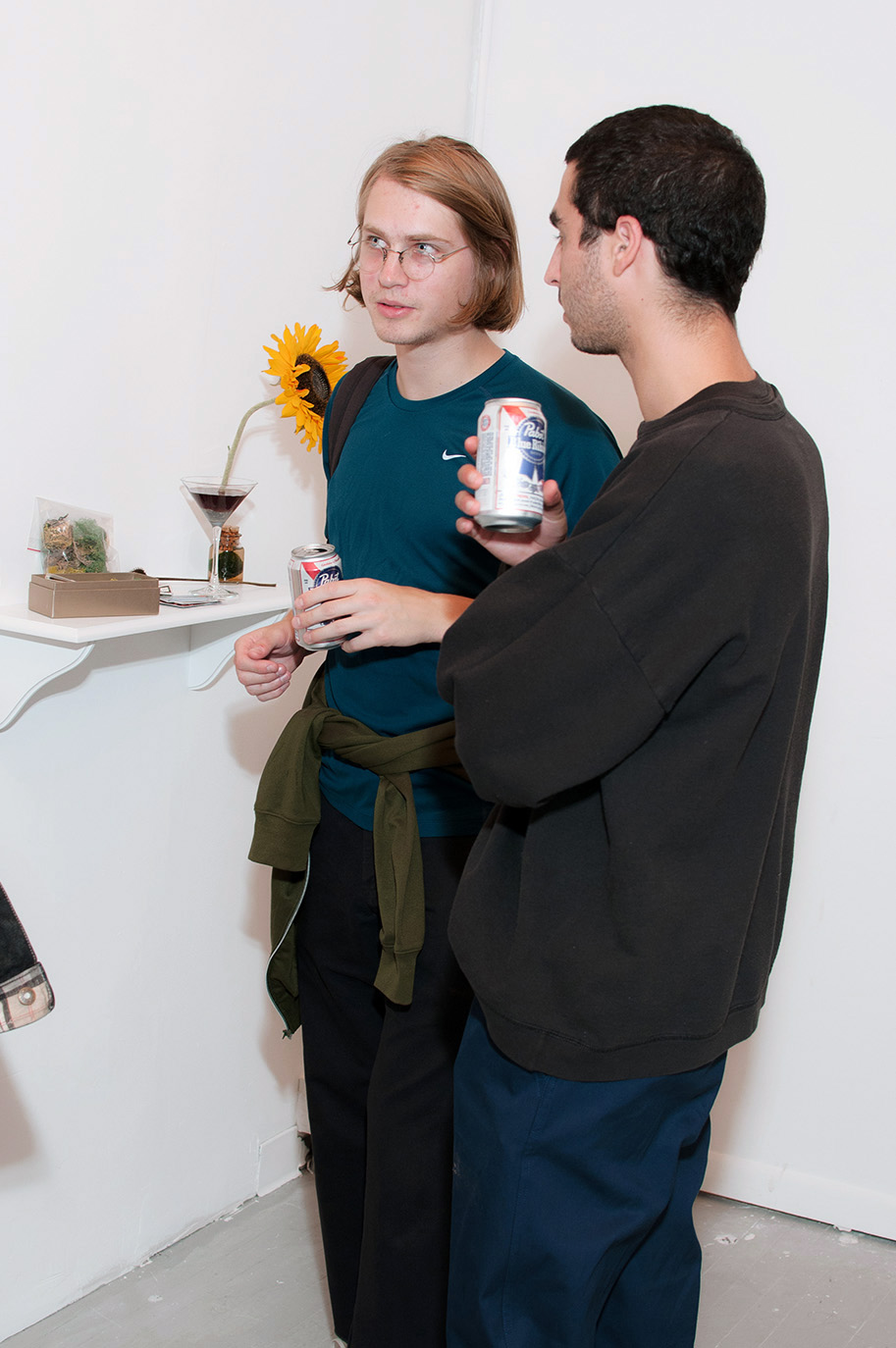 Two young men holding beers and talking next to a shelf with small sculptures